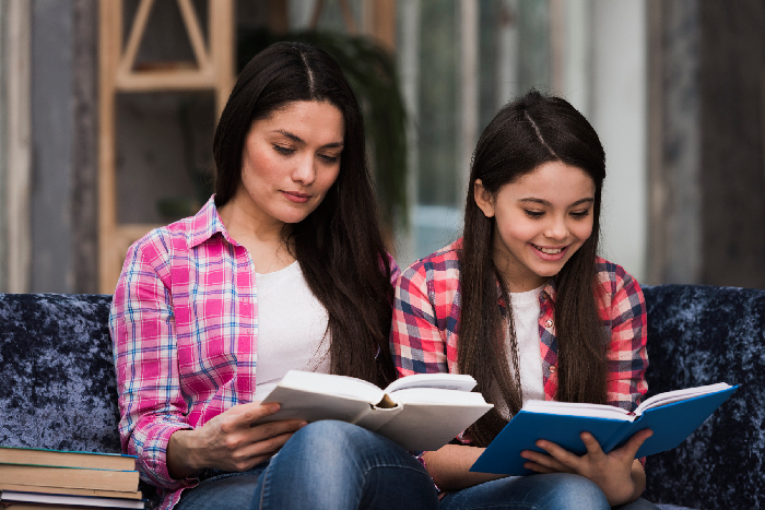 A mother and daughter reading a physical book together happily in a cozy home living room setting.